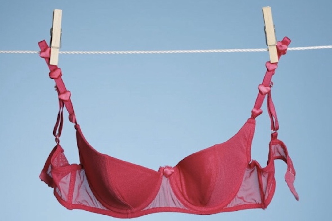 Color photo of magenta colored bra hung up with clothespins with blue sky background.