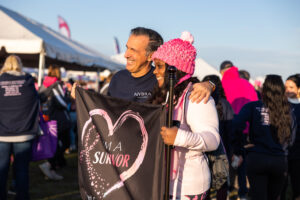 Side angle shot of a man and a woman stand side by side holding a banner that says "I'm a survivor" inside of a pink heart with a breast cancer ribbon.