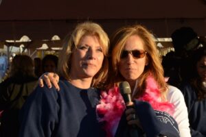 Two women stand shoulder to shoulder at Making Strides of Long Island 2012. The woman on the left wears a blue sweatshirt. The woman on the right has wears sunglasses and a pink feather boa. She is holding a microphone.