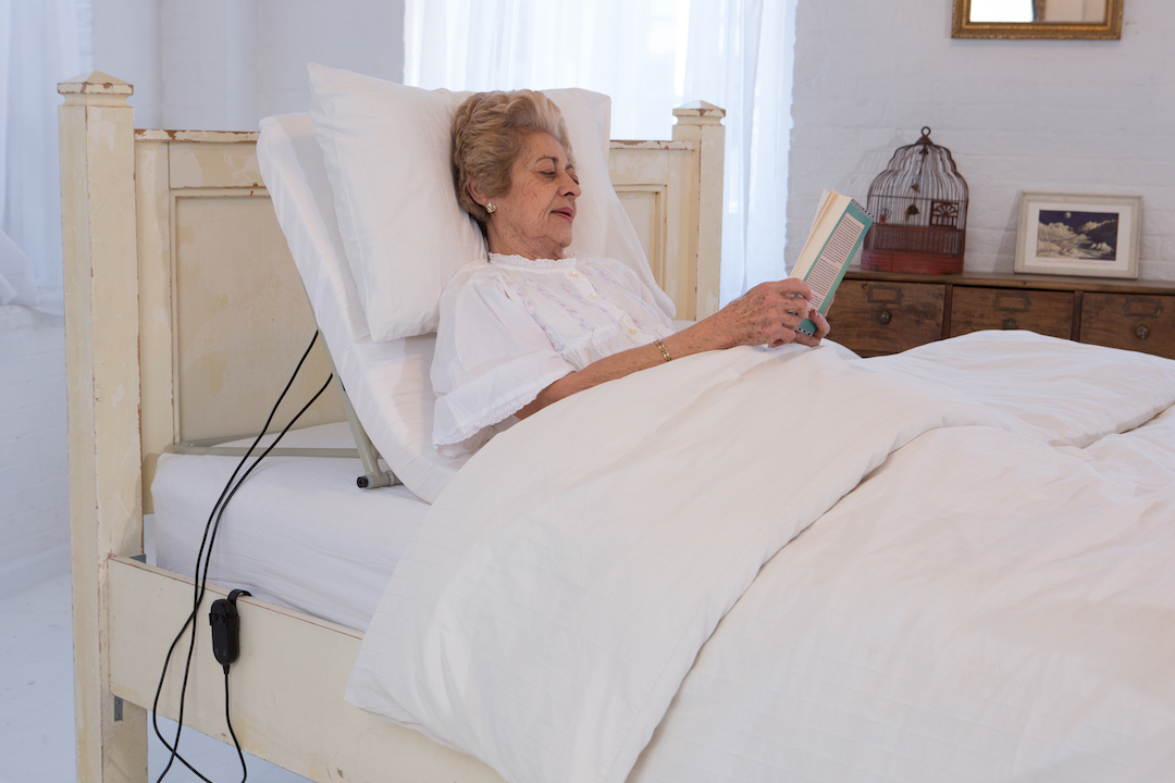 Color photo of woman in bed using the recliner product while she is reading.