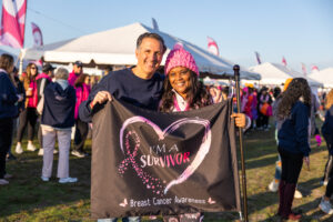 A man and a woman stand side by side holding a banner that says "I'm a survivor" inside of a pink heart with a breast cancer ribbon.