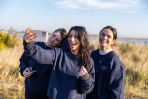 Three women pose for a selfie. The middle woman holds up a cell phone.