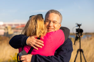 A man and a woman embrace. The man's face is visible over the woman's shoulder. Only the back of her head and back are visible.